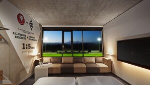 Habitación de hotel con un ventanal grande que da a un campo deportivo, una cama, un sofá y una pared con el logotipo de un equipo deportivo.