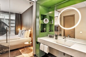 A modern hotel room with a large bed, a green accent wall, a large mirror with a ring light, and a white bathroom vanity. - H4 Hotel Mönchengladbach im BORUSSIA-PARK (Moenchengladbach)