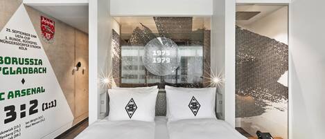 Habitación de hotel con una cama, dos almohadas con un logotipo, una pared con una fecha y texto, y un teléfono en una mesita de noche.