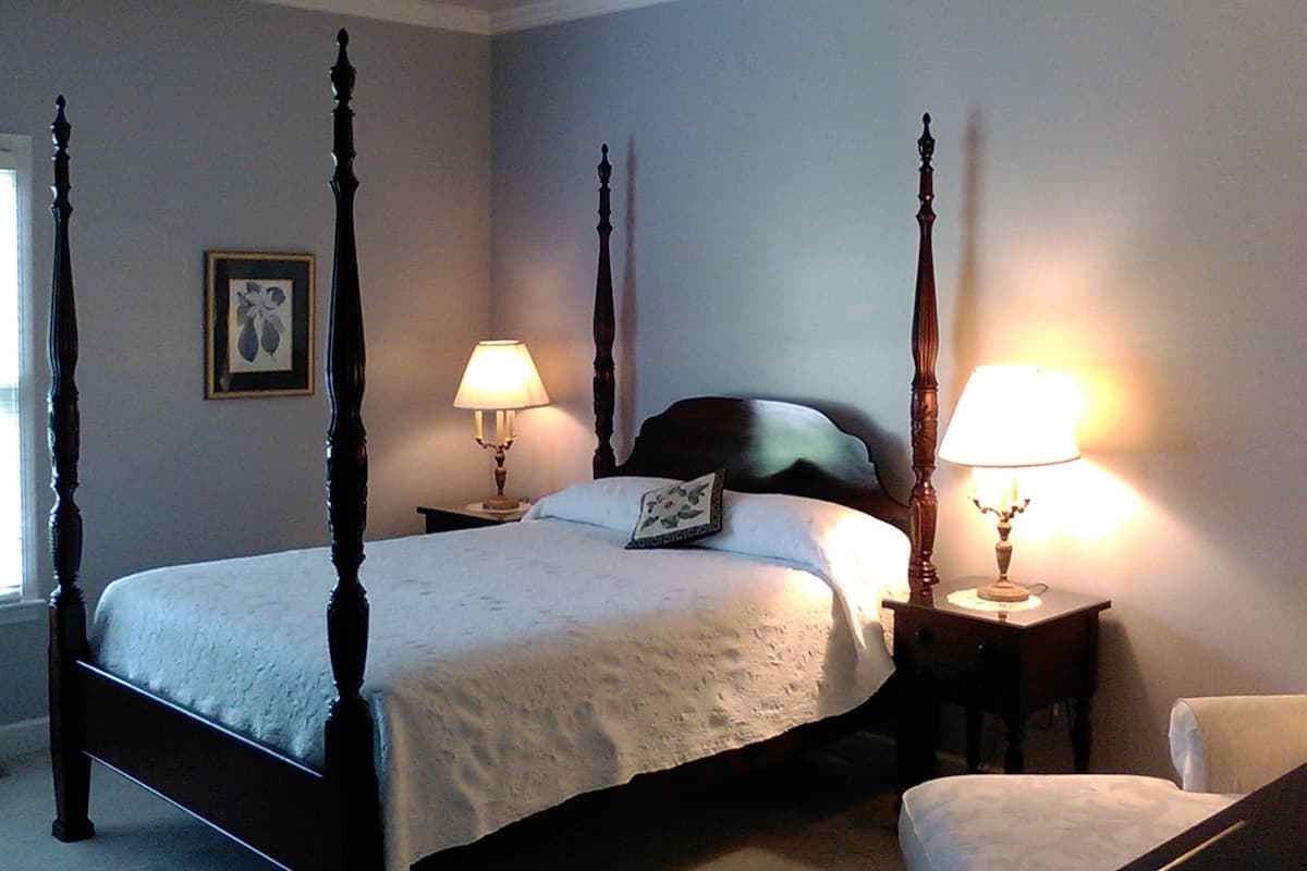 A four-poster bed with a white bedspread, two bedside tables with lamps, a framed picture on the wall, and a chair in the corner.
