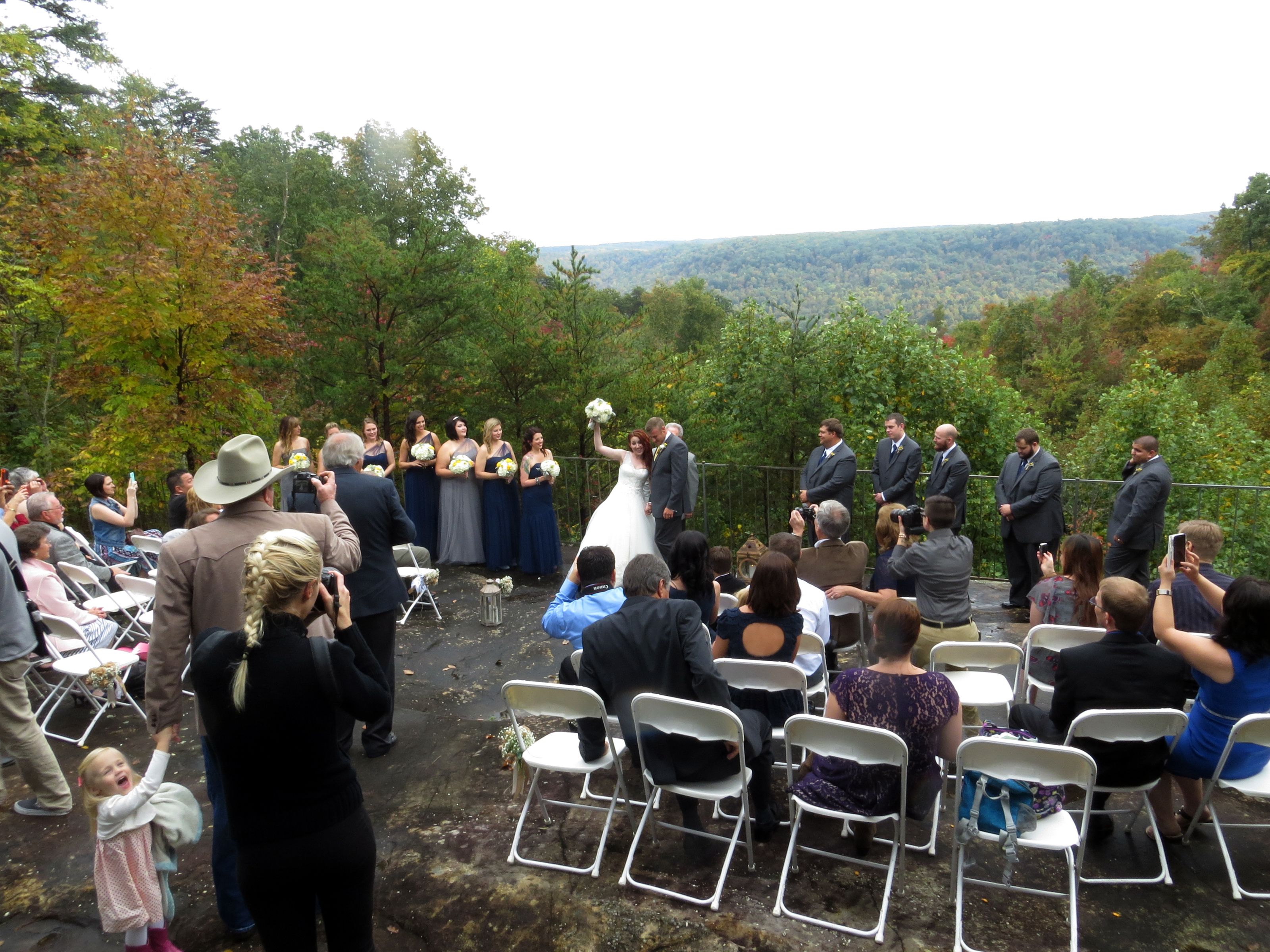 An outdoor wedding ceremony with guests seated on white chairs, surrounded by trees and a mountainous backdrop.