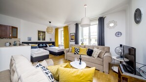 A modern living room with a sofa, a coffee table, and a TV. There is a kitchen area with a sink and cabinets.