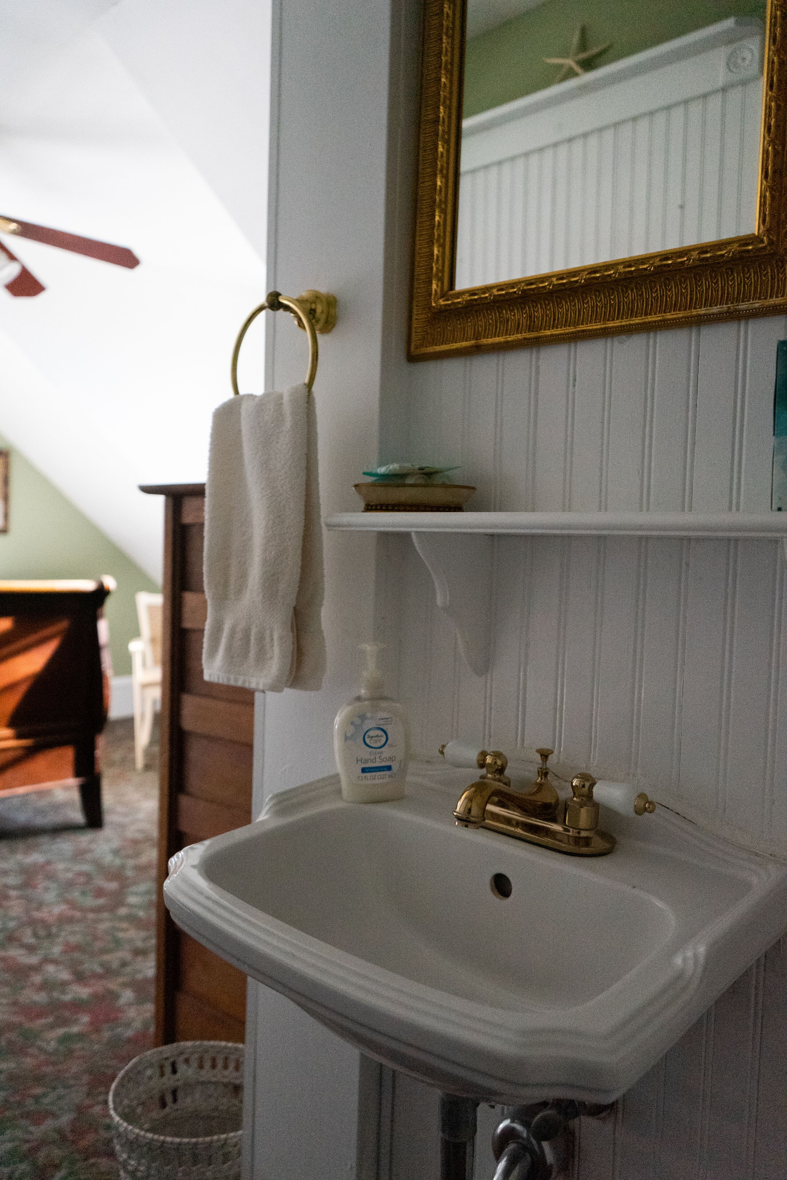Une salle de bain avec un lavabo blanc, des robinets dorés, un miroir encadré d’or et une serviette suspendue à un porte-serviettes doré.