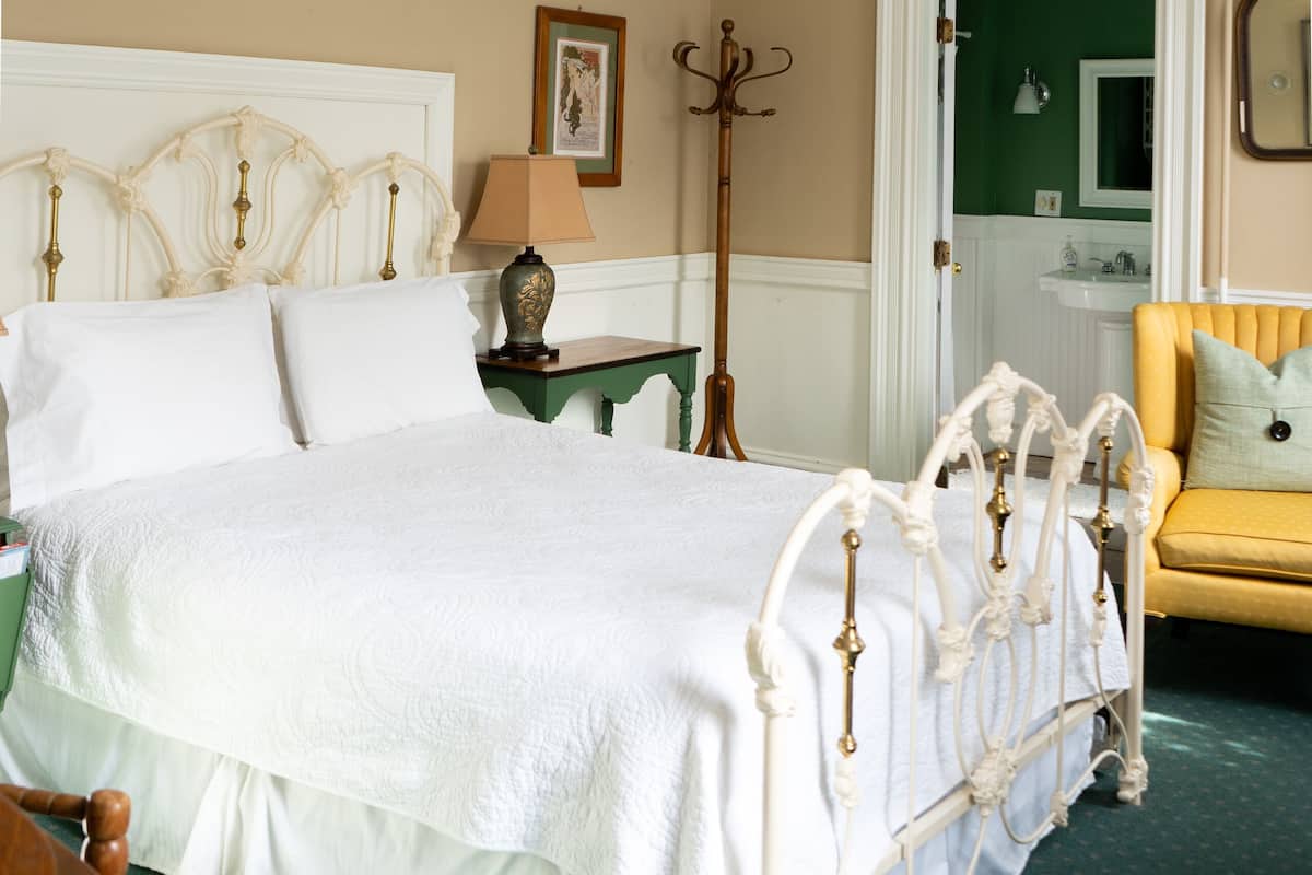 A bedroom with a bed, a nightstand, a lamp, a chair, a ceiling fan, and a door leading to a bathroom.