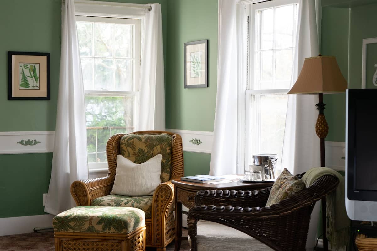 A room with green walls, a bed, two wicker chairs, a small table, a lamp, and a television.