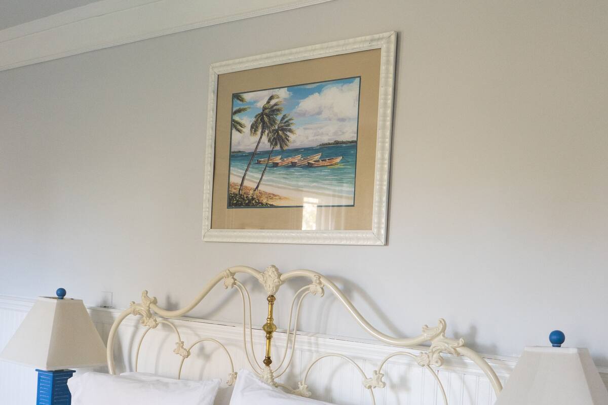 A neatly made bed with white linens, two bedside tables with lamps, and a framed painting of a beach scene on the wall.