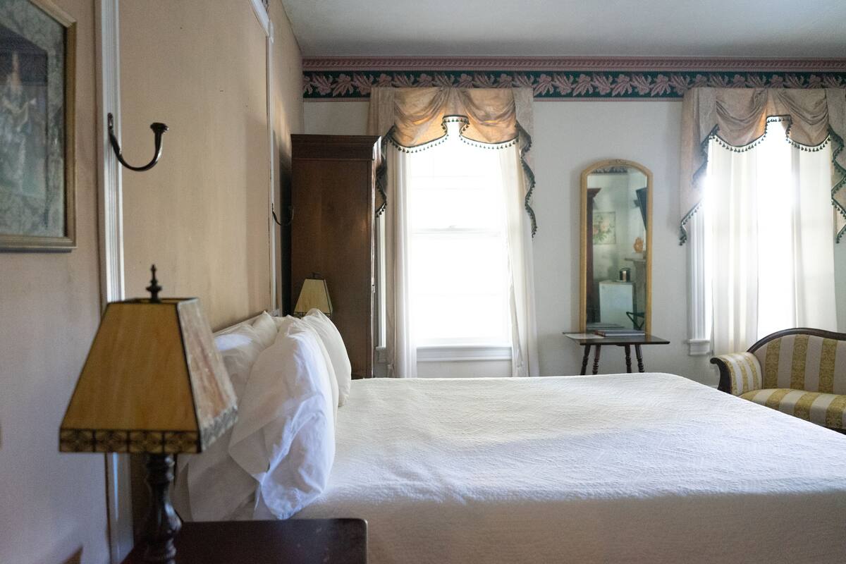 A hotel room with a large bed, a bedside table with a lamp, a chair, a small table, and a mirror on the wall.