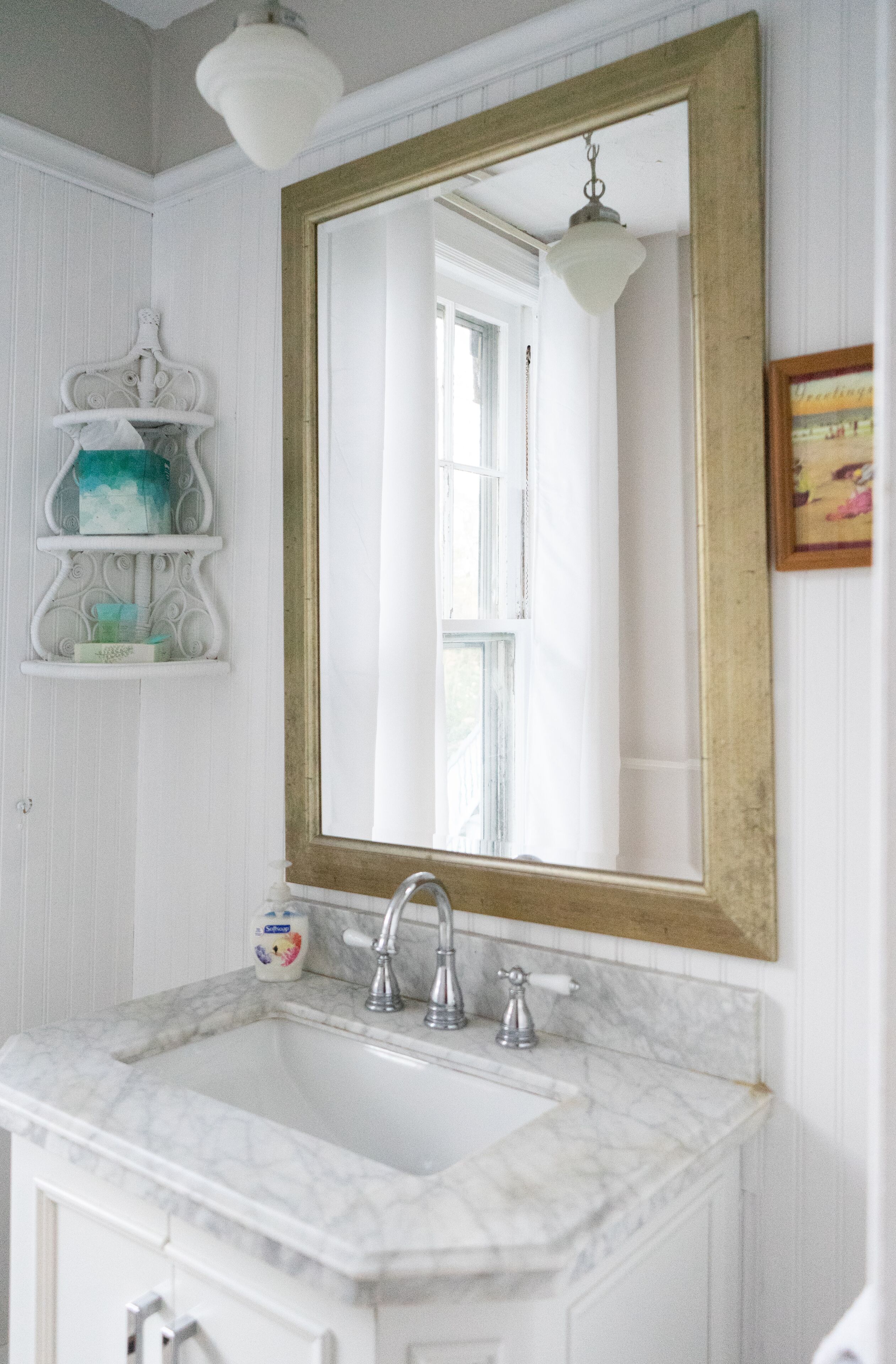 Une salle de bain avec un lavabo en marbre blanc, un miroir encadré d’or et un mur blanc avec une fenêtre.