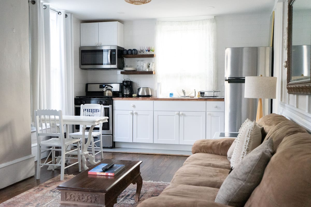 A modern kitchen with white cabinets, a microwave, and a refrigerator. There is a dining area with a white table and chairs. A beige sofa is in the living room area.