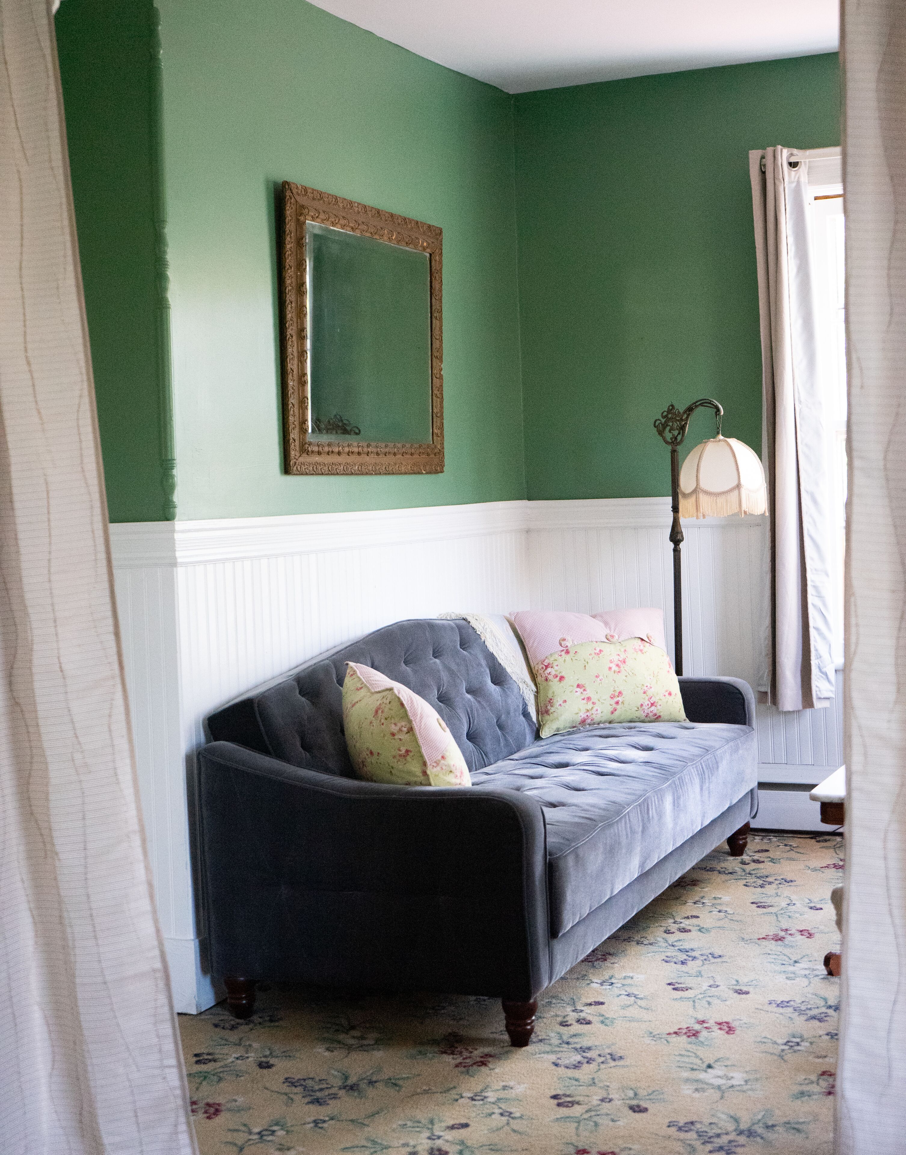 A room with a dark grey sofa, floral patterned carpet, a mirror on the wall, and a floor lamp.