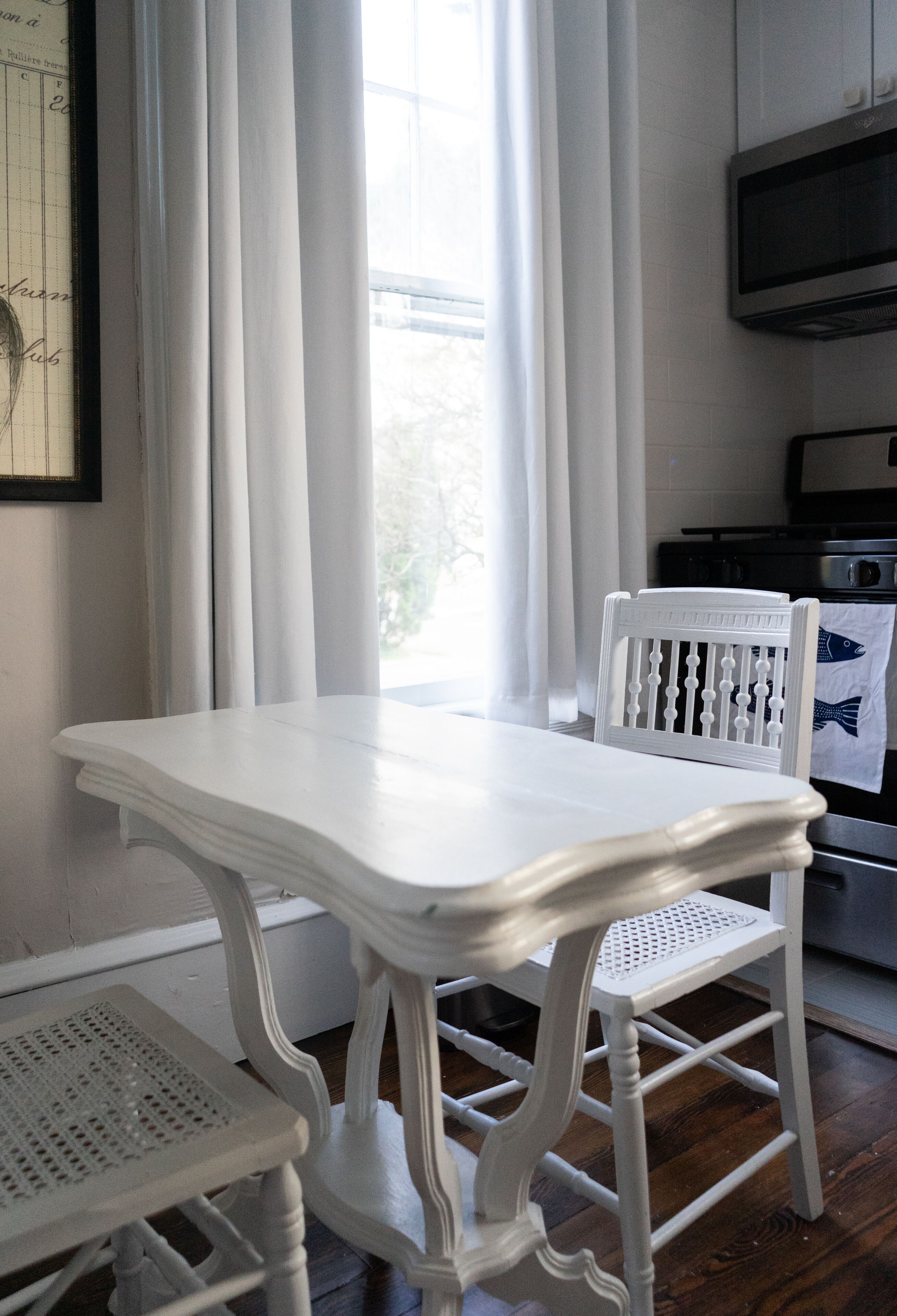 Une table à manger blanche avec des chaises, une fenêtre avec des rideaux et un coin cuisine équipé d’un four à micro-ondes et d’un four traditionnel.