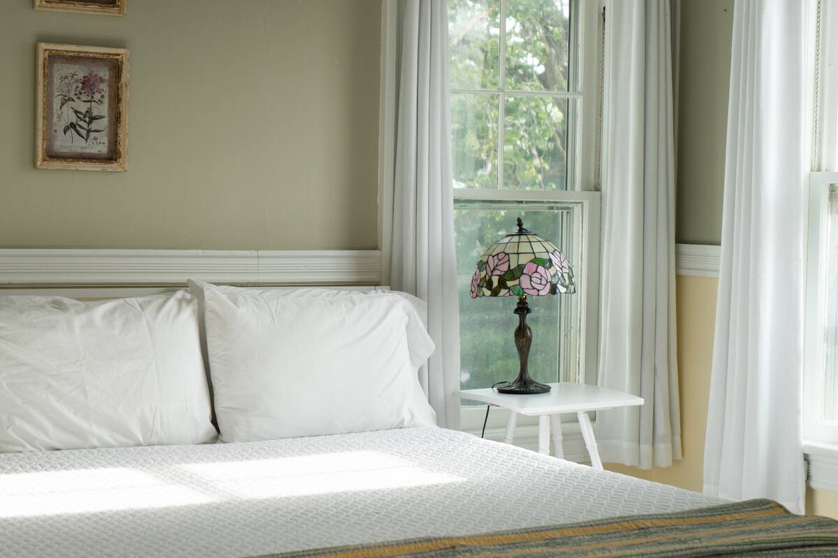 A neatly made bed with white linens, a small bedside table with a lamp, and a window with curtains.