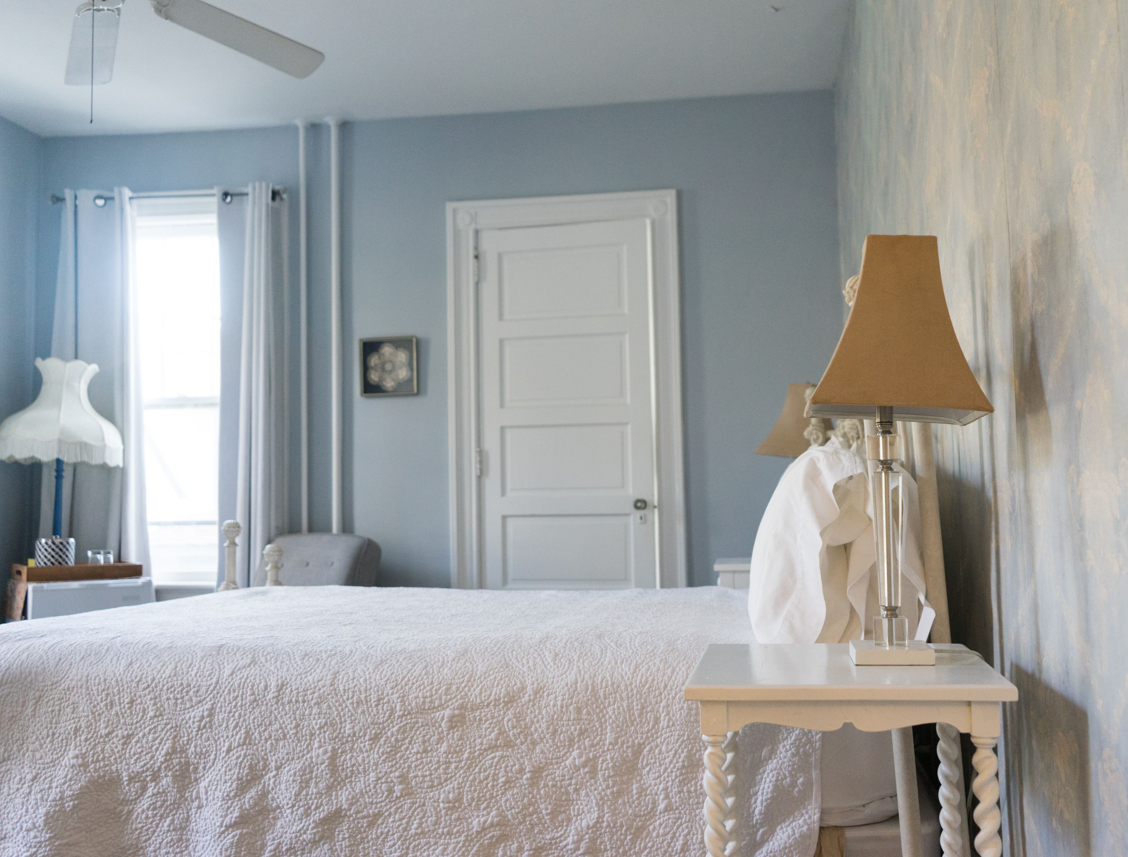 Un dormitorio con cama, mesita de noche, lámpara, puerta y ventana con cortinas.