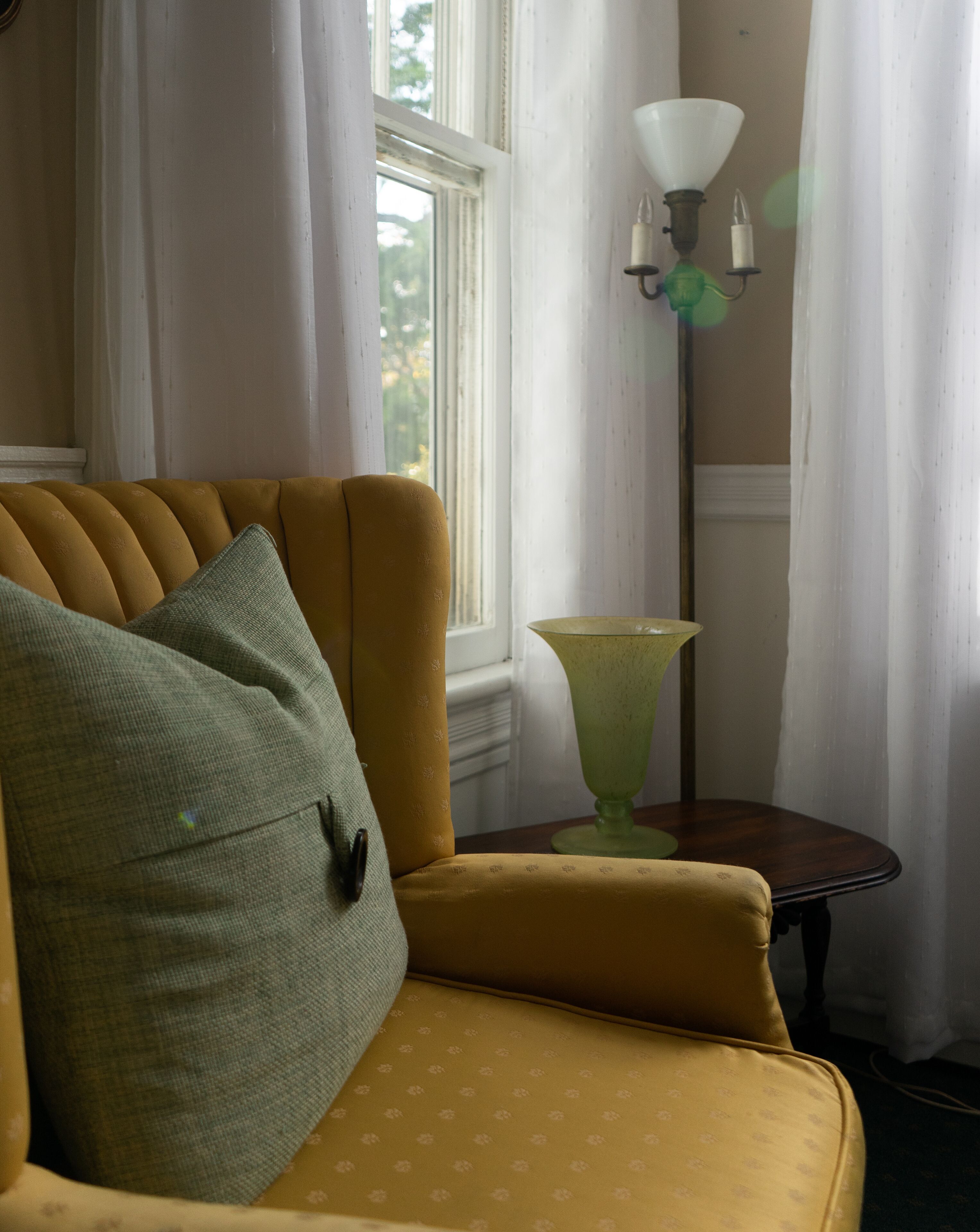 A yellow armchair with a green pillow, a green floor lamp, and a window with white curtains.