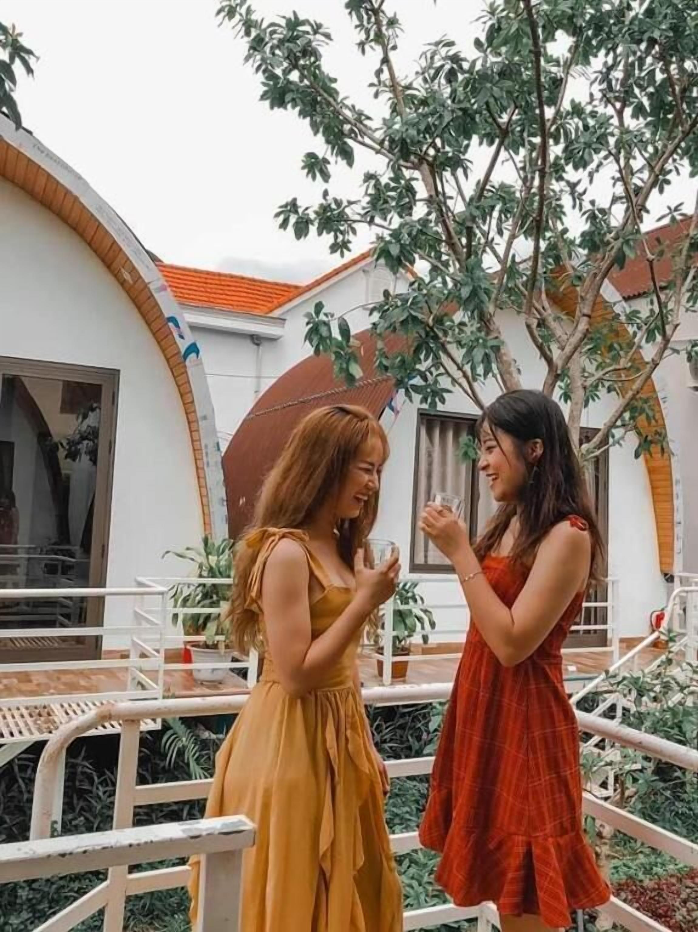 Two women conversing on a balcony with a white building and a tree in the background.