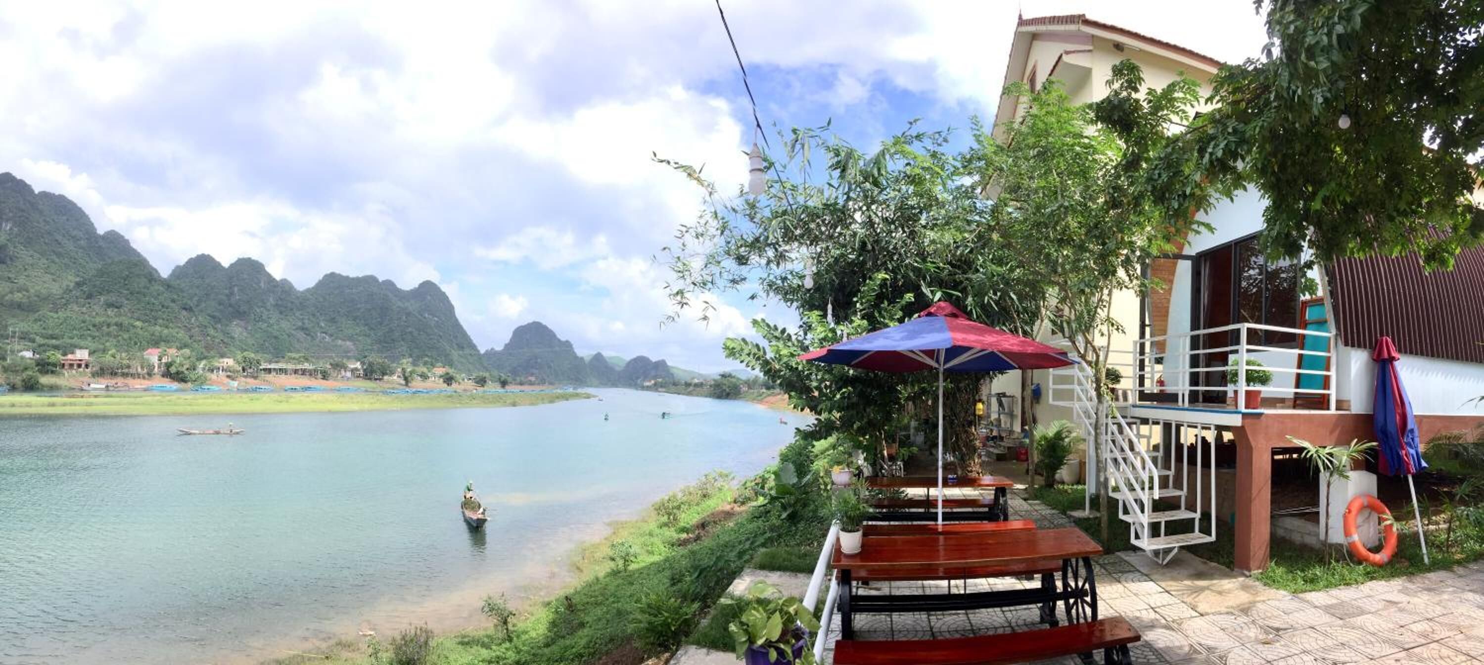A riverside view with a boat, a dining area with tables and an umbrella, and a building with a balcony.