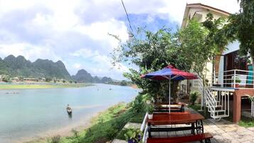 A riverside view with a boat, a dining area with tables and an umbrella, and a building with a balcony.