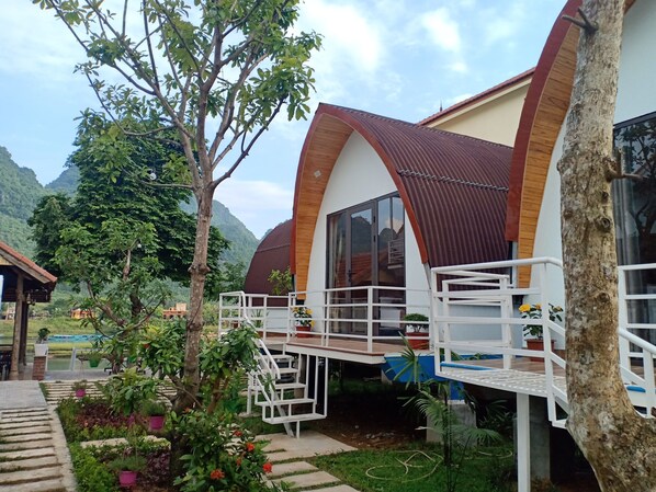 A modern building with a curved roof and large windows, surrounded by lush greenery and mountains in the background. - Areca Bungalow (Bo Trach)