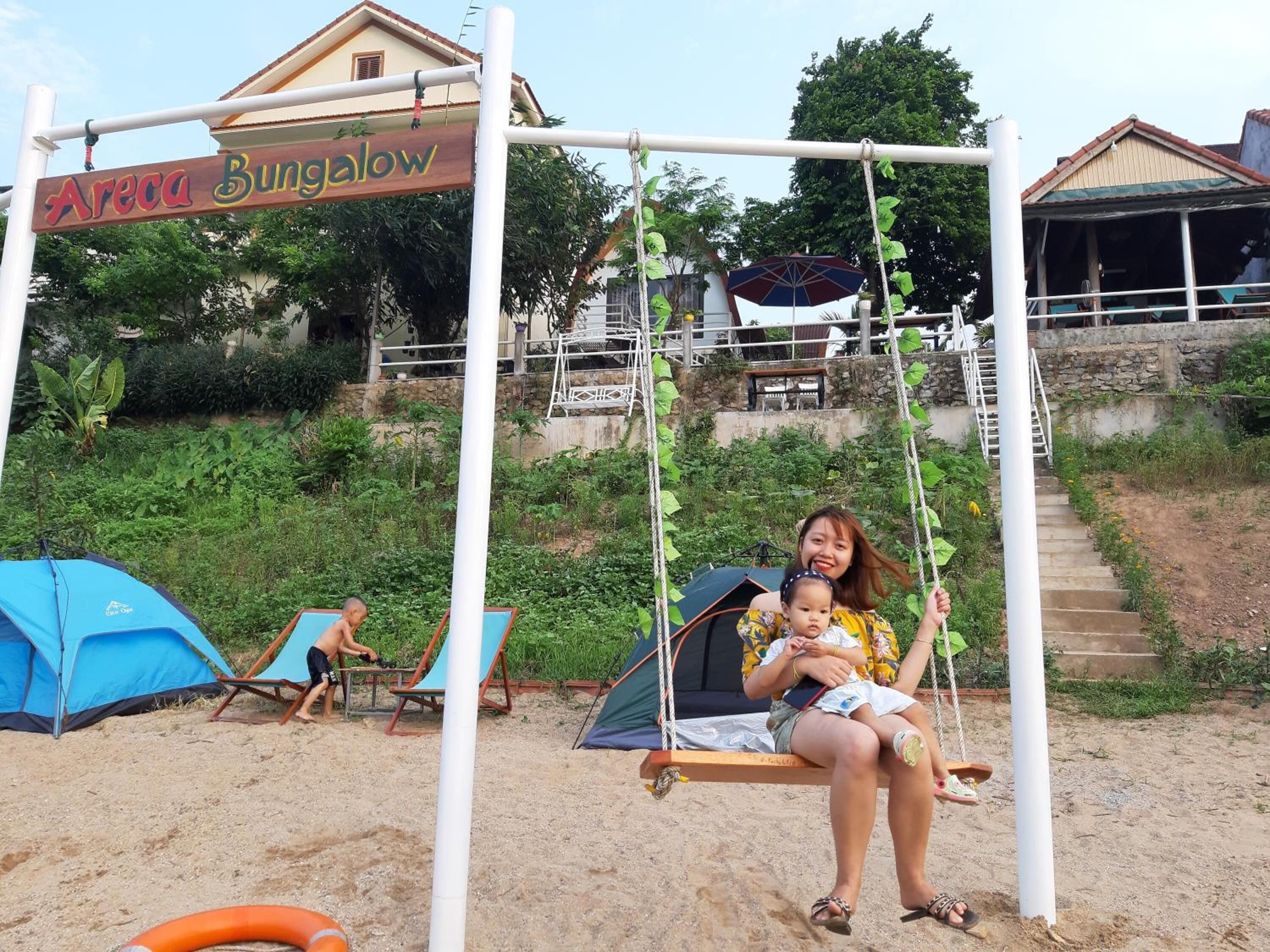 A swing set labeled "Areca Bungalow" with a person and a child on it, tents, and a staircase leading to a building.
