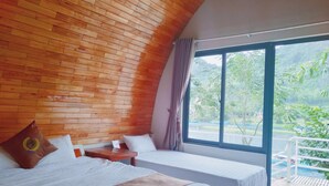 A room with a wooden ceiling, two beds, a window with a view of greenery, and a pool area. - Areca Bungalow (Phong Nha)