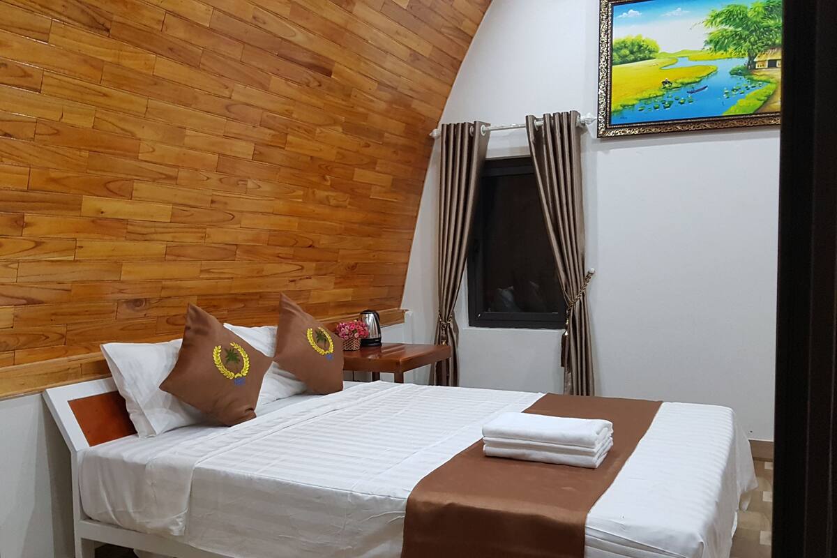 A neatly made bed with white linens and a brown accent, a wooden headboard, a nightstand with a kettle and flowers, a window with curtains, and a painting on the wall.