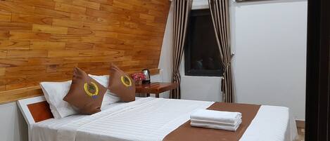 A neatly made bed with white linens and a brown accent, a wooden headboard, a nightstand with a kettle and flowers, a window with curtains, and a painting on the wall.
