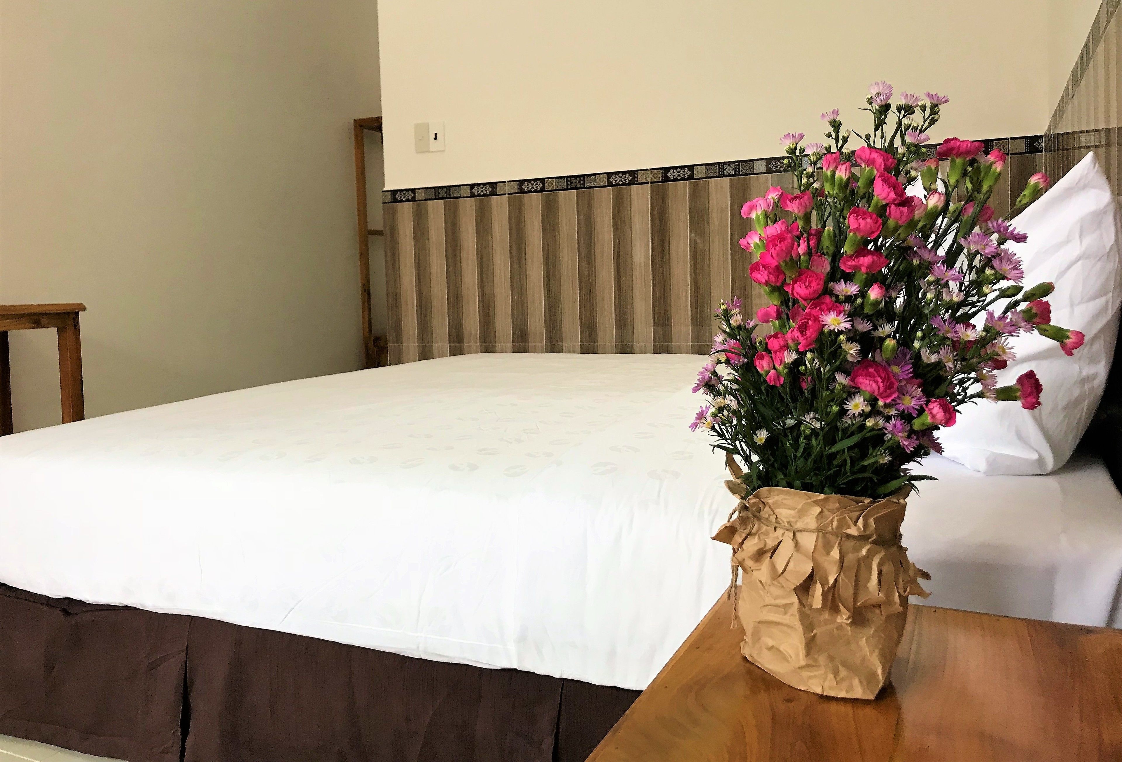 Ein ordentlich bezogenes Bett mit weißer Bettdecke und einem Holzkopfstück. Auf einem Nachttisch neben dem Bett liegt eine braune Papiertüte mit einem Strauß aus rosa und violetten Blumen.