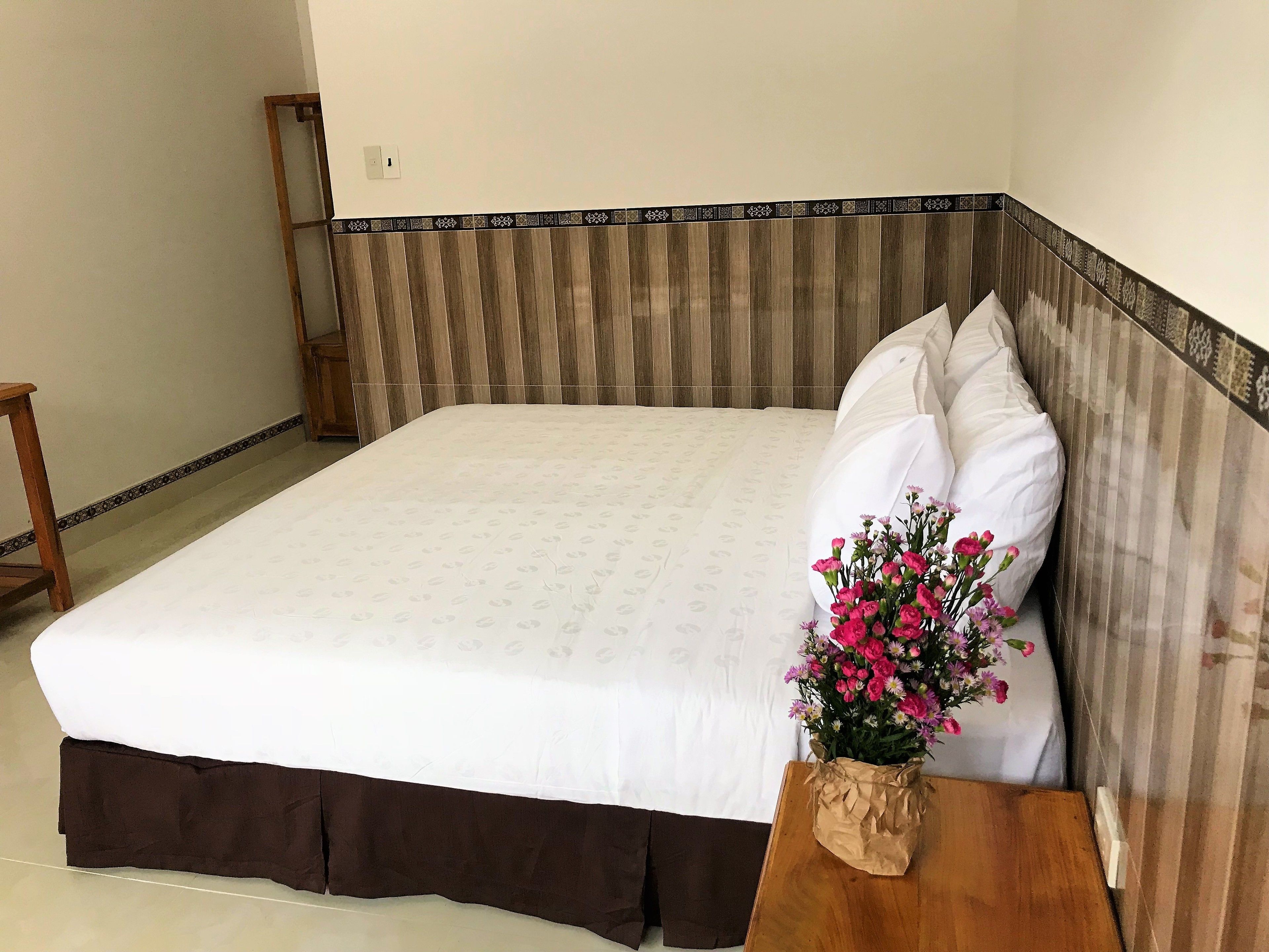 A neatly made bed with a patterned bedspread, a wooden nightstand with a vase of flowers, and a wooden headboard with a decorative border.