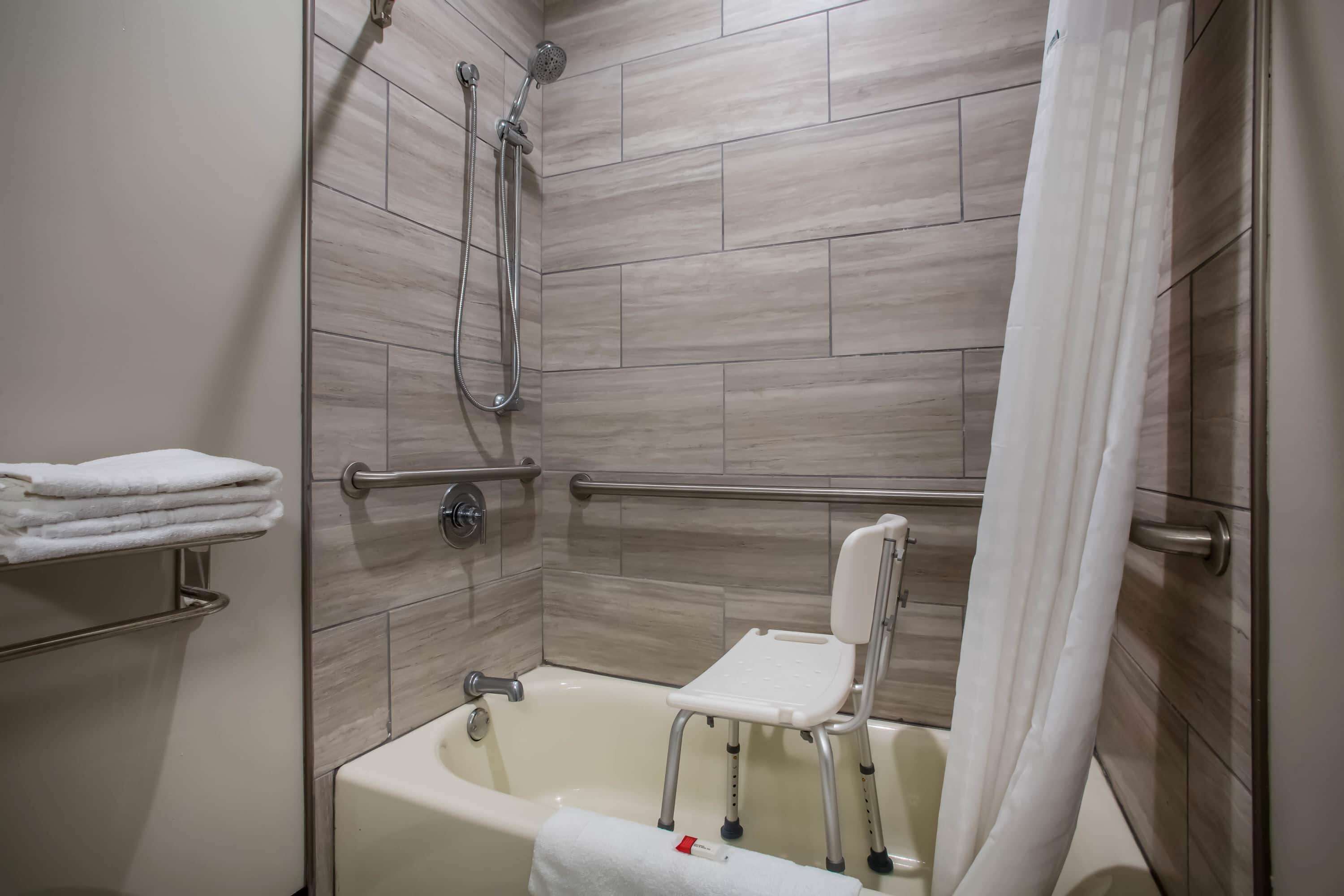 Un área de ducha con barra de apoyo, una silla blanca y un perchero para toallas.