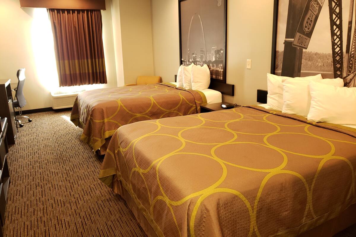 Une chambre d’hôtel avec deux lits, un bureau, une chaise, une fenêtre avec des rideaux et une affiche représentant un paysage urbain accrochée au mur.