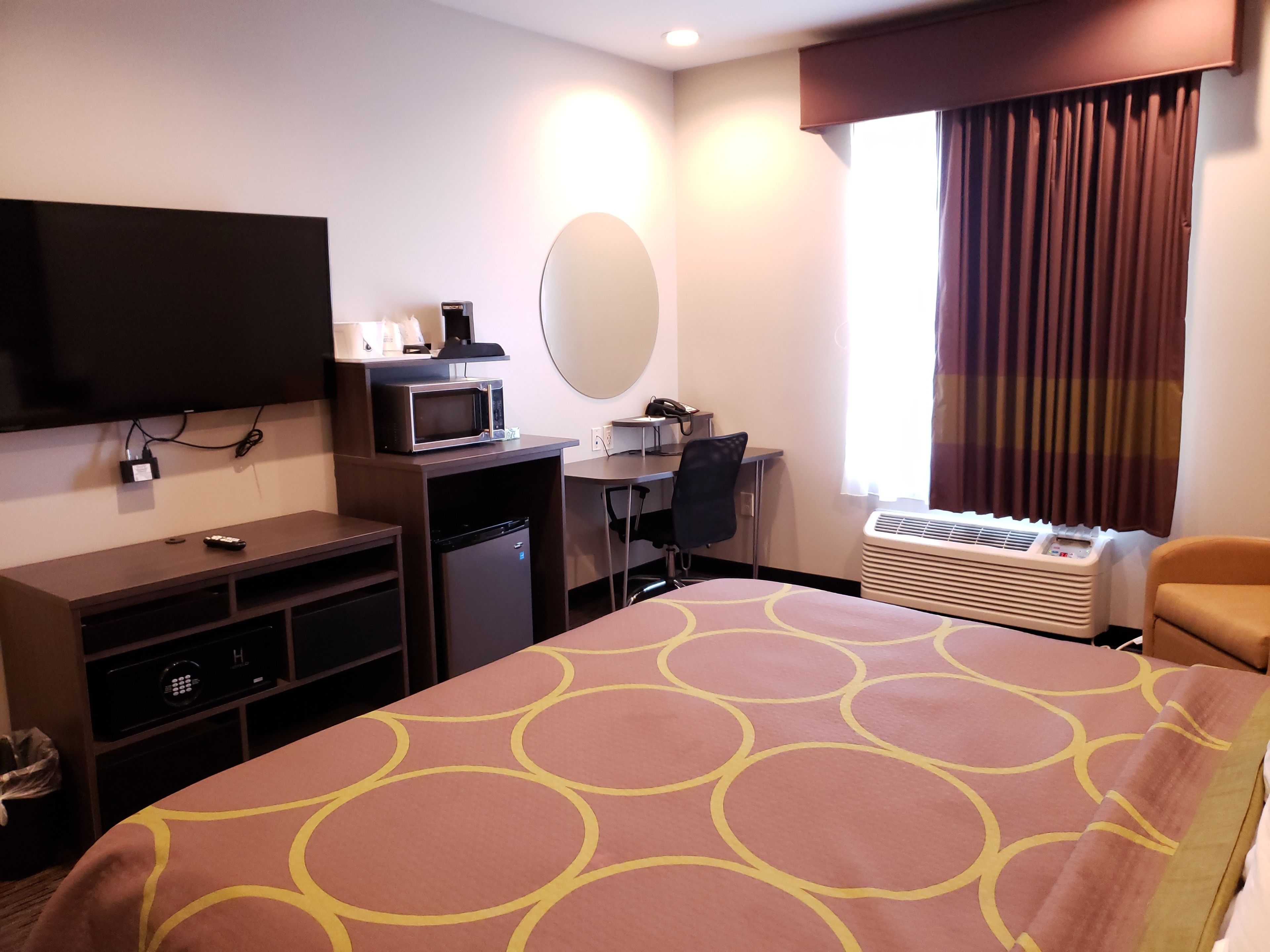A hotel room with a bed, a desk, a chair, a television, a microwave, a heater, and a window with curtains.