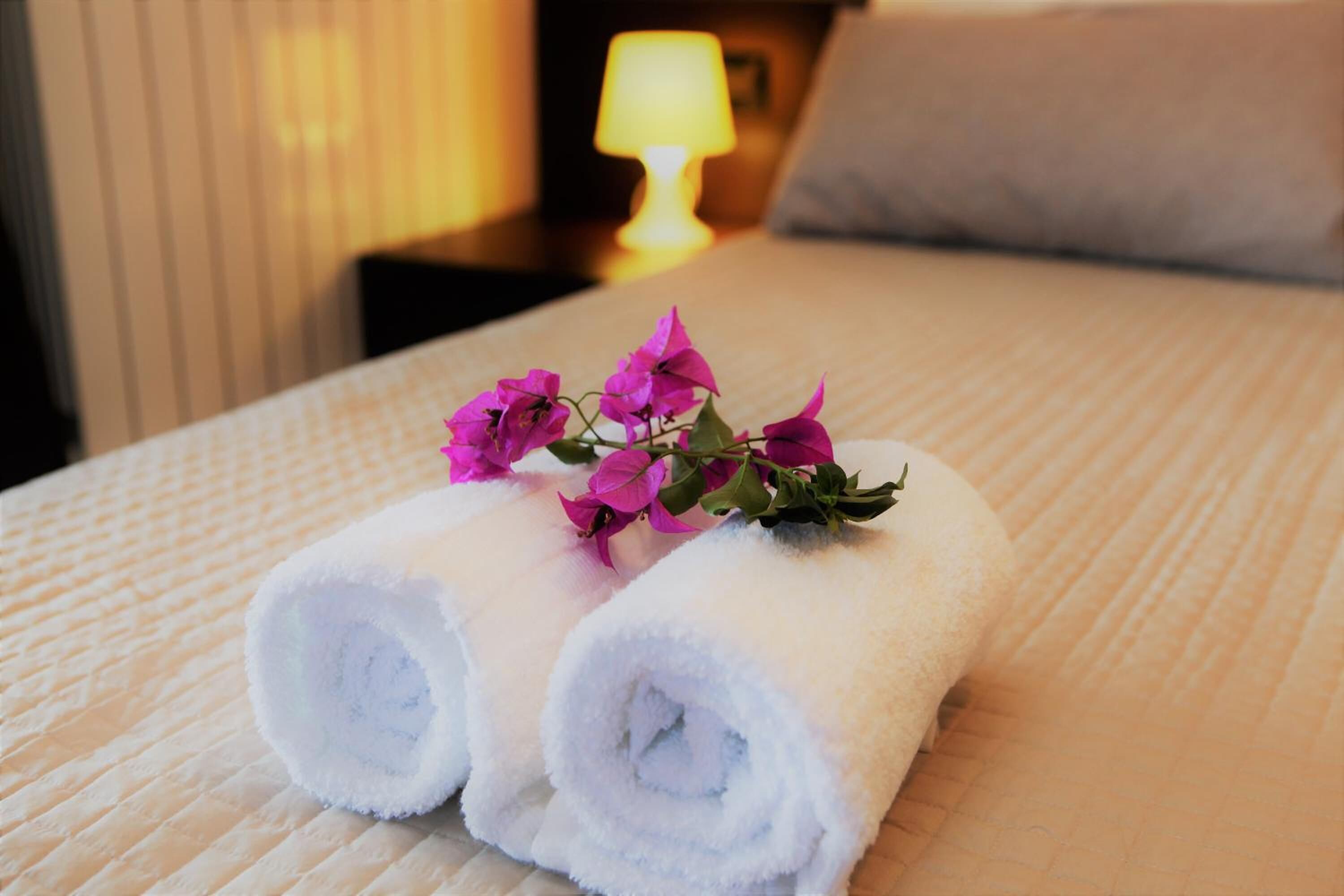 Un letto rifatto con due asciugamani bianchi arrotolati e un piccolo bouquet di fiori rosa sopra.