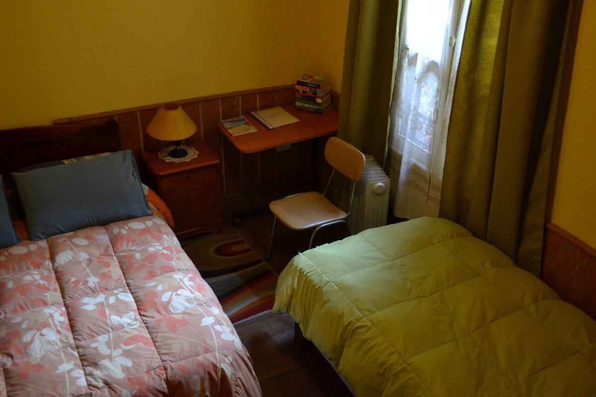 Una habitación de hotel pequeña con dos camas, un escritorio con lámpara, una silla y un radiador.