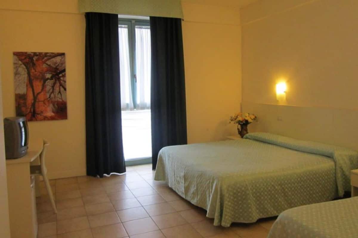 A hotel room with two beds, a desk, a chair, a painting, a window with curtains, and a wall-mounted light.