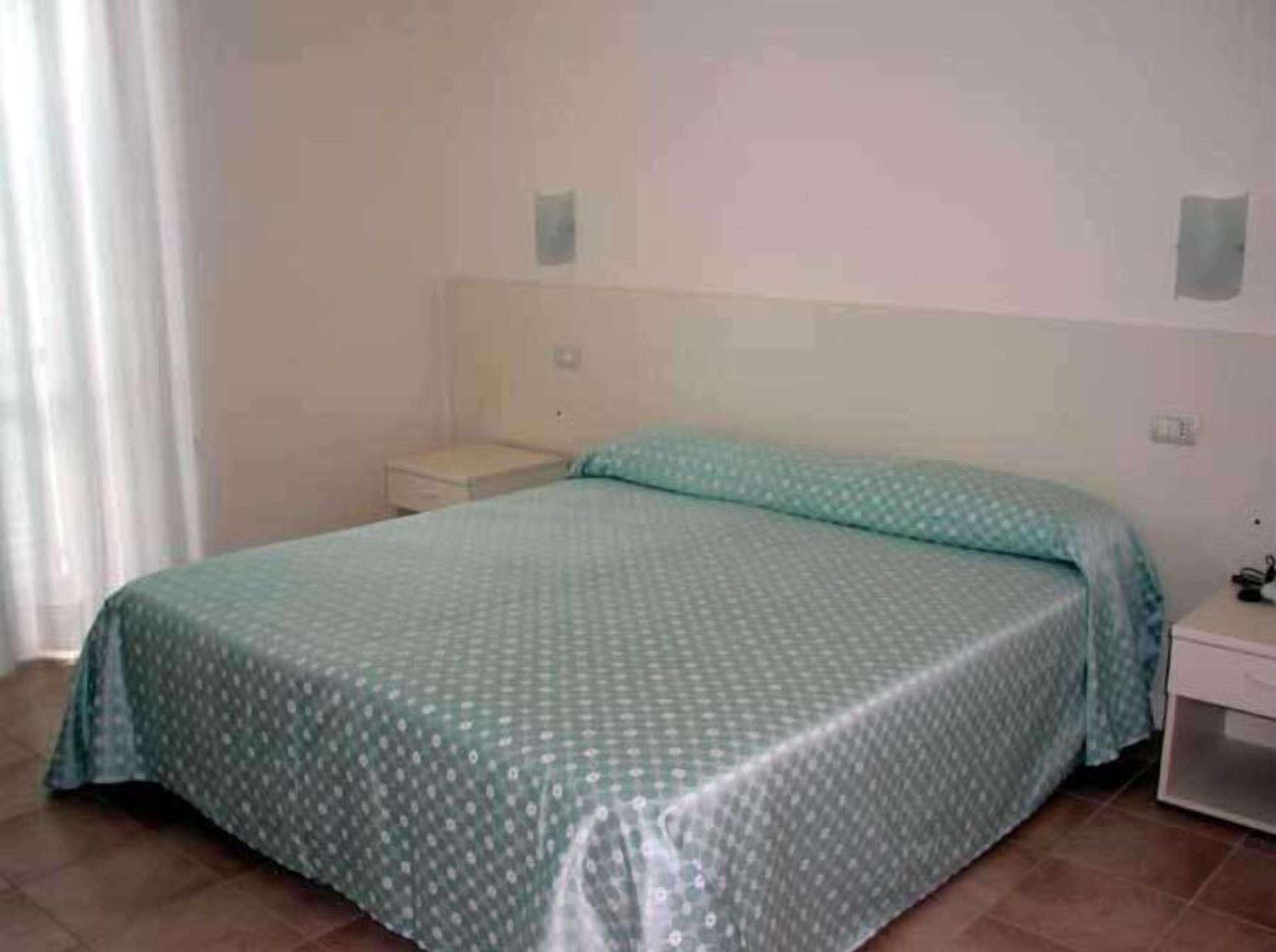 Une chambre avec un lit simple, une couvre-lit à motifs verts, une petite table de chevet et un mur blanc uni.