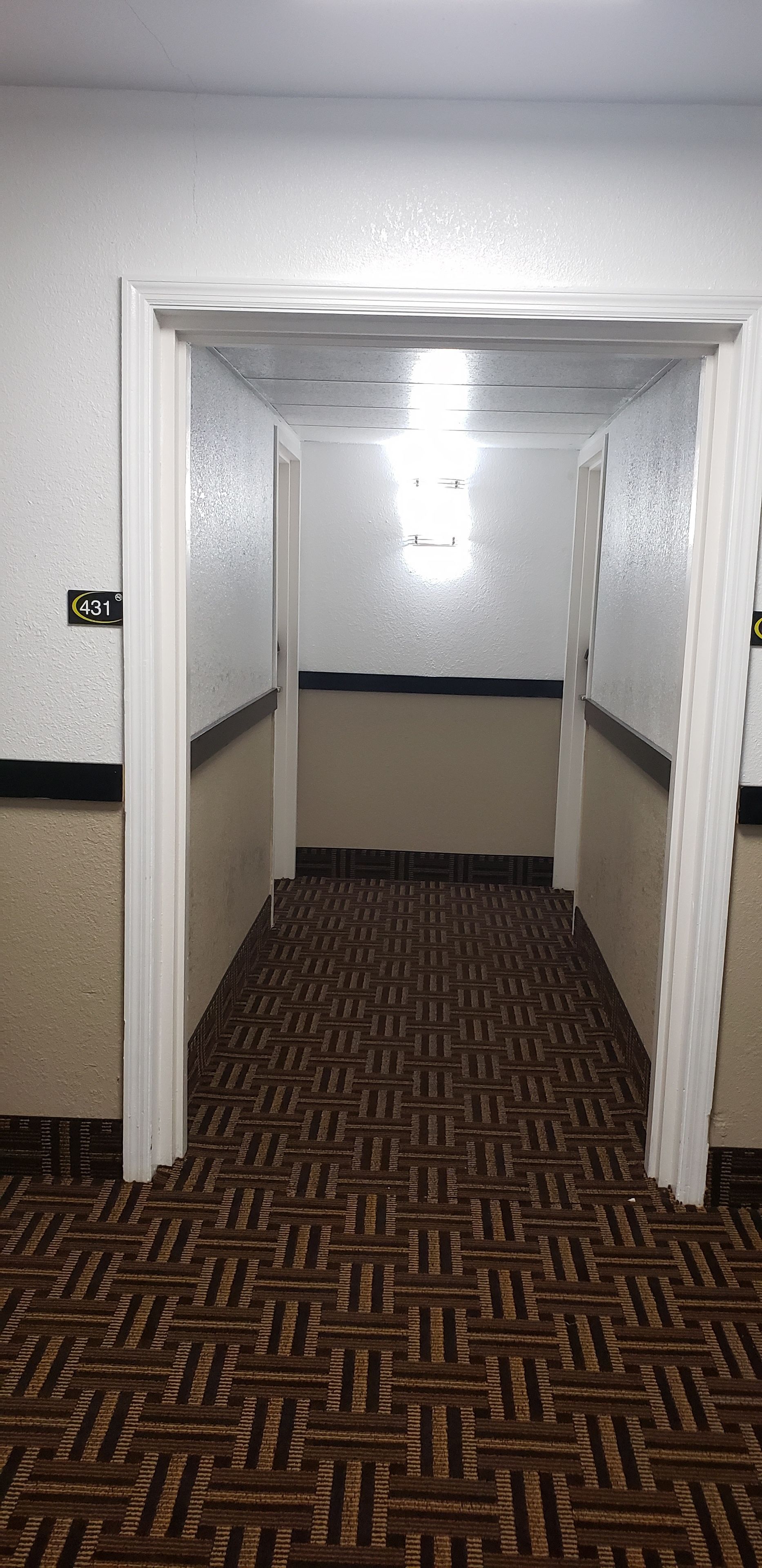 A narrow hallway with patterned carpet, white walls, and a door with a number sign.