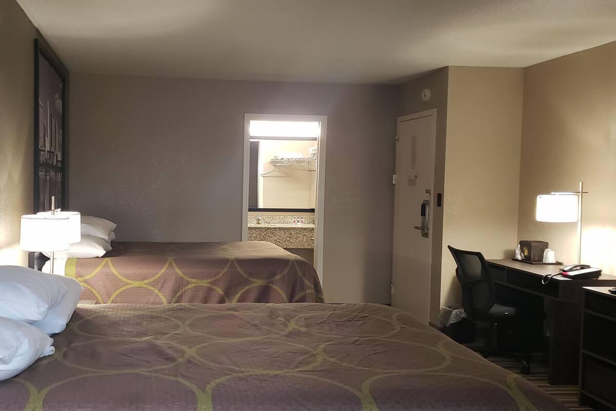 A hotel room with two beds, a desk, and a chair. There is a window with a view of buildings, a door leading to a bathroom, and a wall with a cityscape poster.