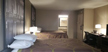 Habitación de hotel con dos camas, un escritorio y una silla. Se ve una ventana con vistas a edificios, una puerta que da al baño y un cartel con un paisaje urbano en la pared.