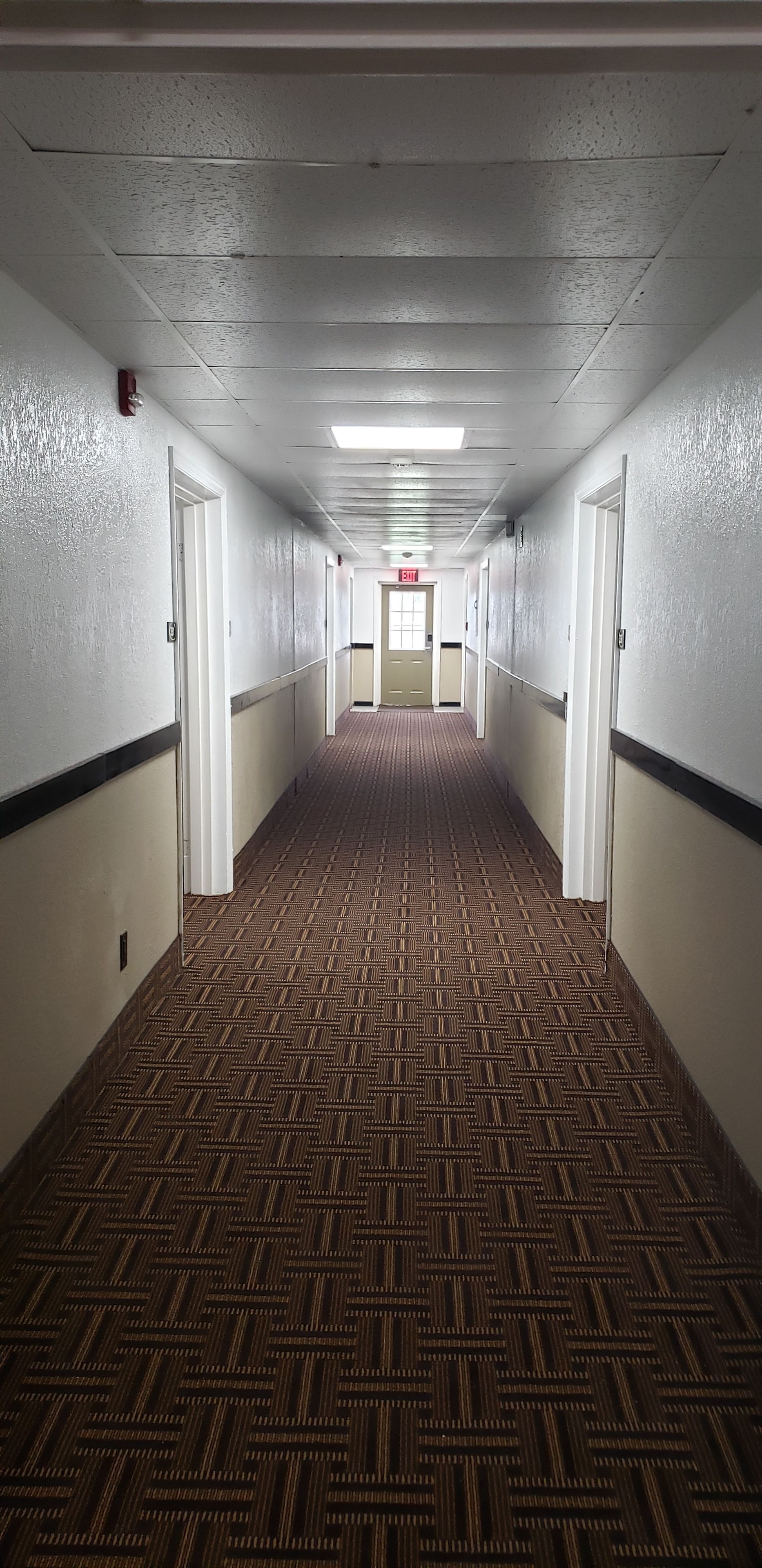 A long, narrow hotel corridor with patterned carpet, white walls, and doors on both sides.