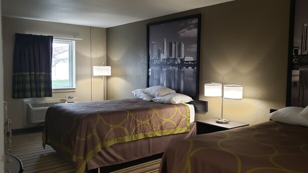 A hotel room with two beds, a window with curtains, a wall-mounted air conditioner, and a large cityscape painting. - Travel Inn Of Sidney Ohio (Sidney)