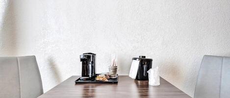 Un espacio comedor limpio y minimalista, con una mesa de madera, dos cafeteras negras, una cesta pequeña con utensilios y un salero y pimentero.