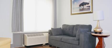 Una habitación con un sofá gris, una mesita redonda con lámpara, un radiador y una ventana con cortinas.