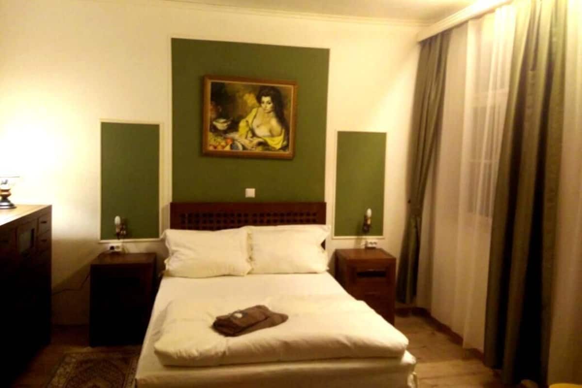 Hotelkamer met een bed, nachtkastje, lamp, schilderij aan de muur en raam met gordijnen.