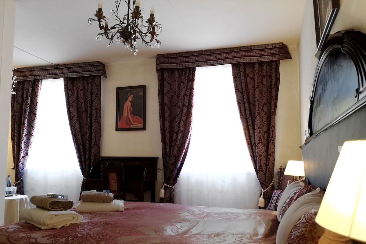 Een hotelkamer met een bed, twee raamgorden, een kroonluchter, een schilderij en een nachtkastje met een lamp.