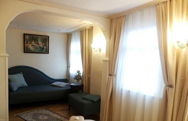 A room with a dark-colored sofa, a small ottoman, a painting on the wall, and a window with curtains.
