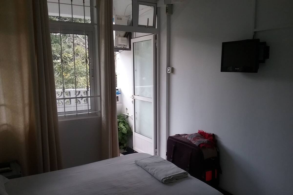 Een hotelkamer met een bed, een raam met gordijnen, een klein tafeltje met een rood voorwerp en een aan de muur bevestigde televisie.