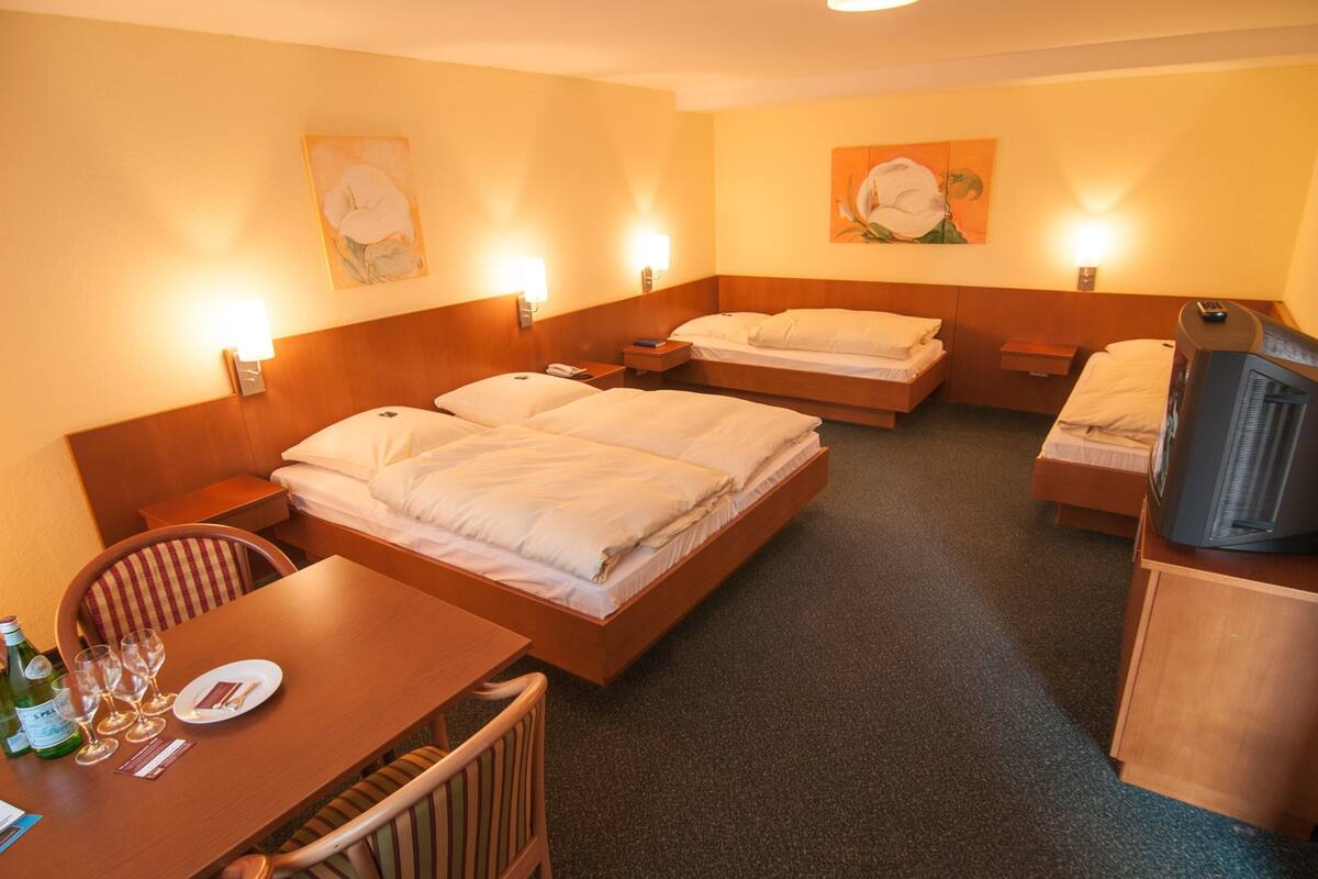 Quarto de hotel com duas camas, televisão, mesa de jantar com copos e garrafa, e obras de arte nas paredes.