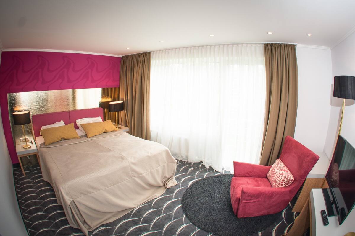 Quarto de hotel com uma cama grande, uma poltrona vermelha, uma televisão e uma janela ampla com cortinas.