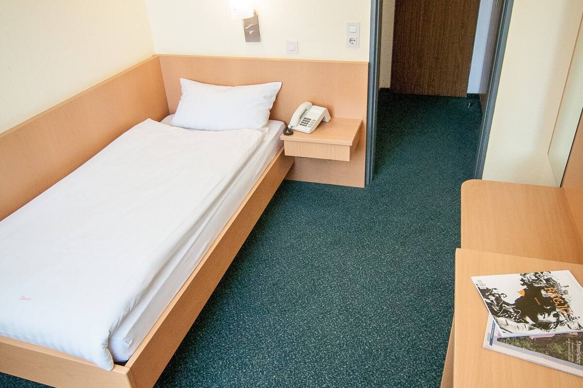 Quarto de hotel com uma cama de solteiro, mesa de cabeceira com telefone, uma cadeira e escrivaninha com controle remoto.