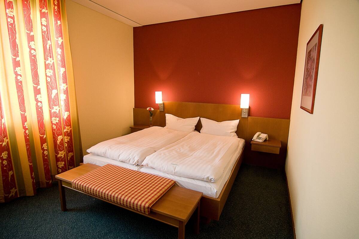 Quarto de hotel com uma cama de casal, mesas de cabeceira com luminárias, um banco e um telefone.